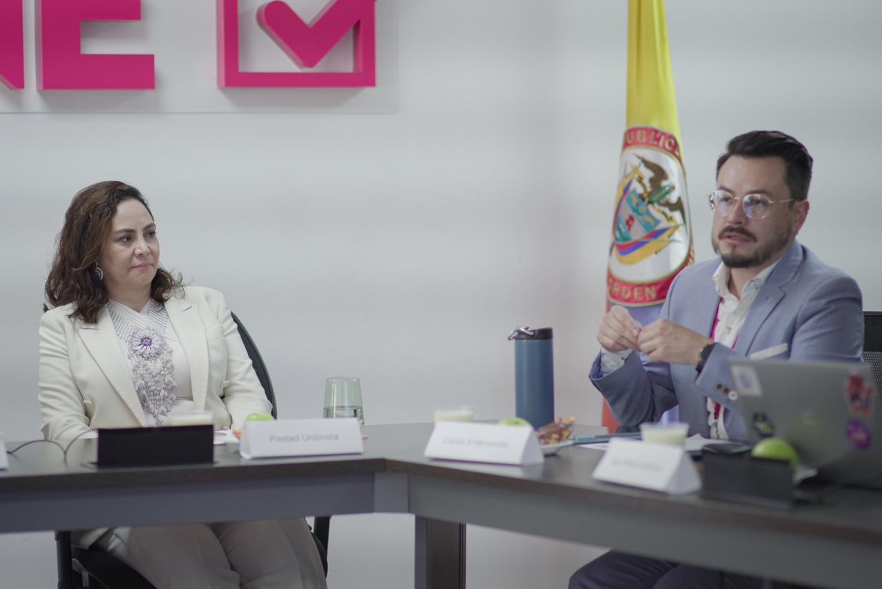 Dos personas, un hombre y una mujer, se sientan a una mesa en una sala de reuniones, con una bandera colombiana y un logotipo de la marca "DANE" en la pared