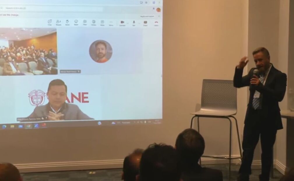 Conferencista realizando una presentación ante un auditorio, mientras en una pantalla se observa una videoconferencia con participantes remotos y una intervención asociada al DANE.