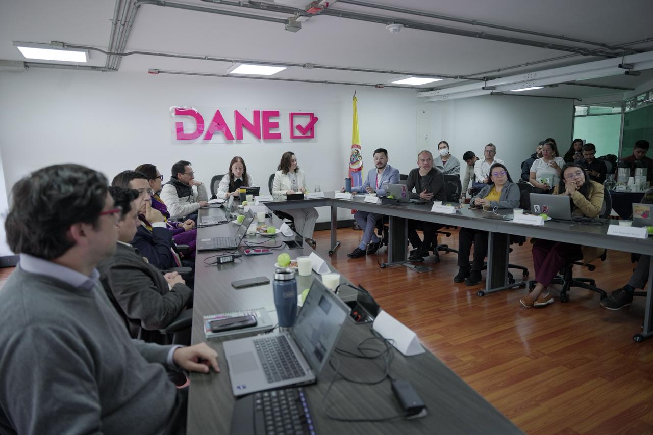 Una fotografía documental muestra a un grupo de personas reunidas alrededor de una mesa en una sala de conferencias, con el logo "DANE" visible en la pared.