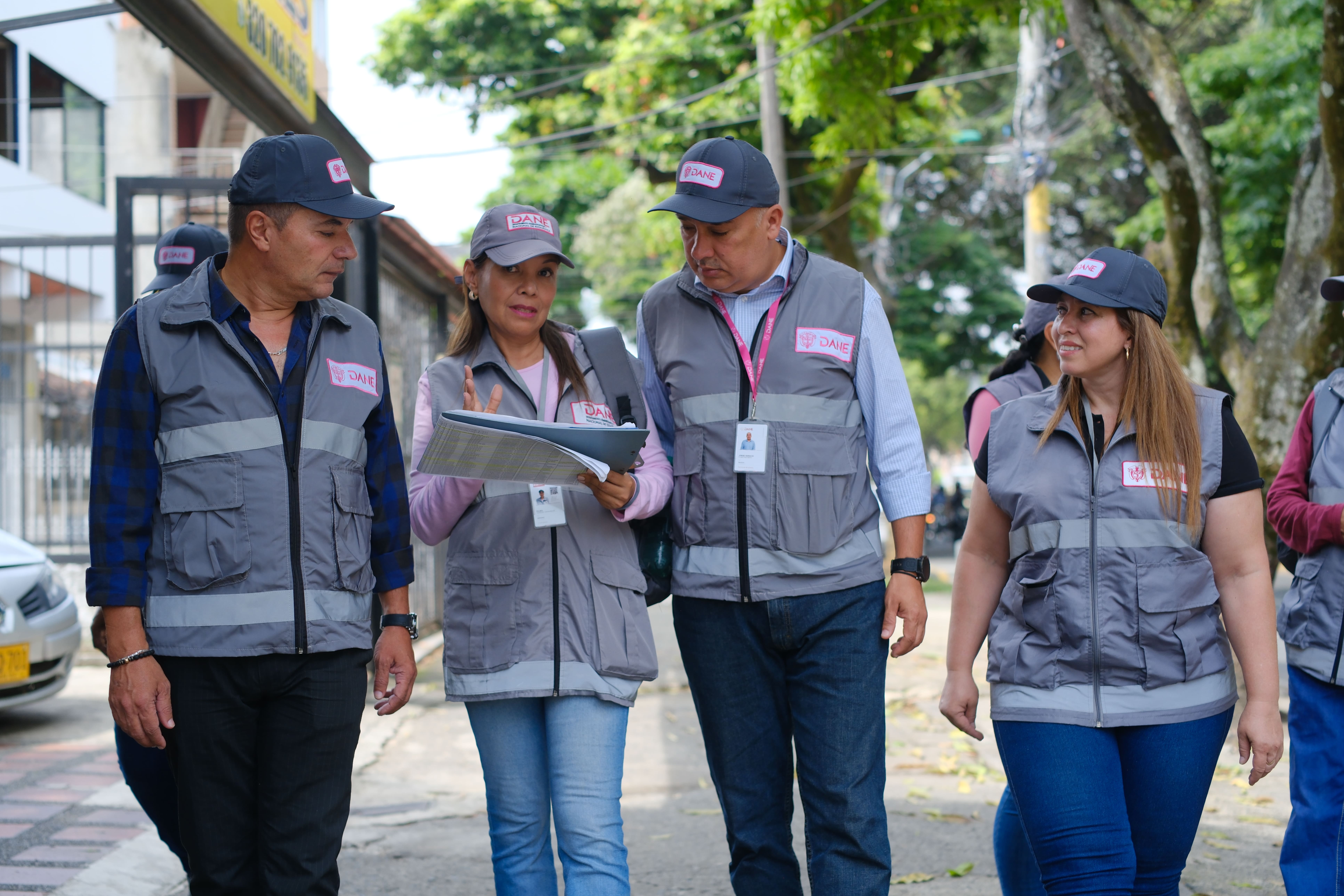 Grupo de cuatro funcionarios del DANE, uniformados con carnet, chaleco y cachucha institucional, caminando juntos por una calle mientras dos de ellos revisan un documento.
