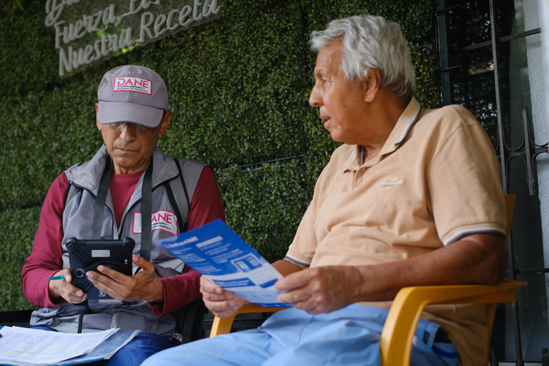 Censista del DANE, con cachucha y chaleco institucional, registrando datos en una tableta junto a un adulto mayor que revisa un folleto