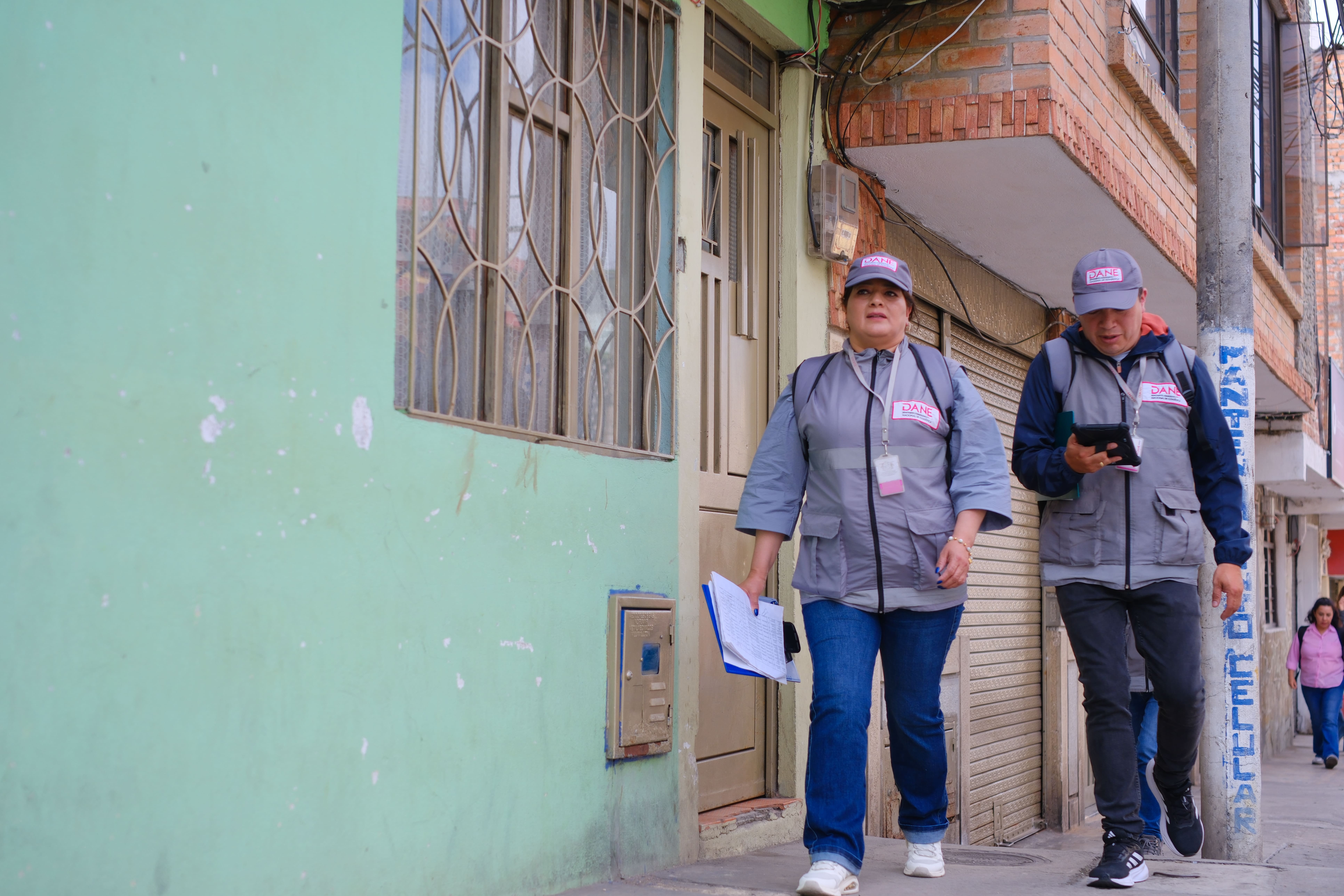 Dos censistas del DANE, uniformados con chaleco y cachucha institucional, caminando por una acera junto a una pared verde; uno sostiene una tableta y la otra una carpeta.
