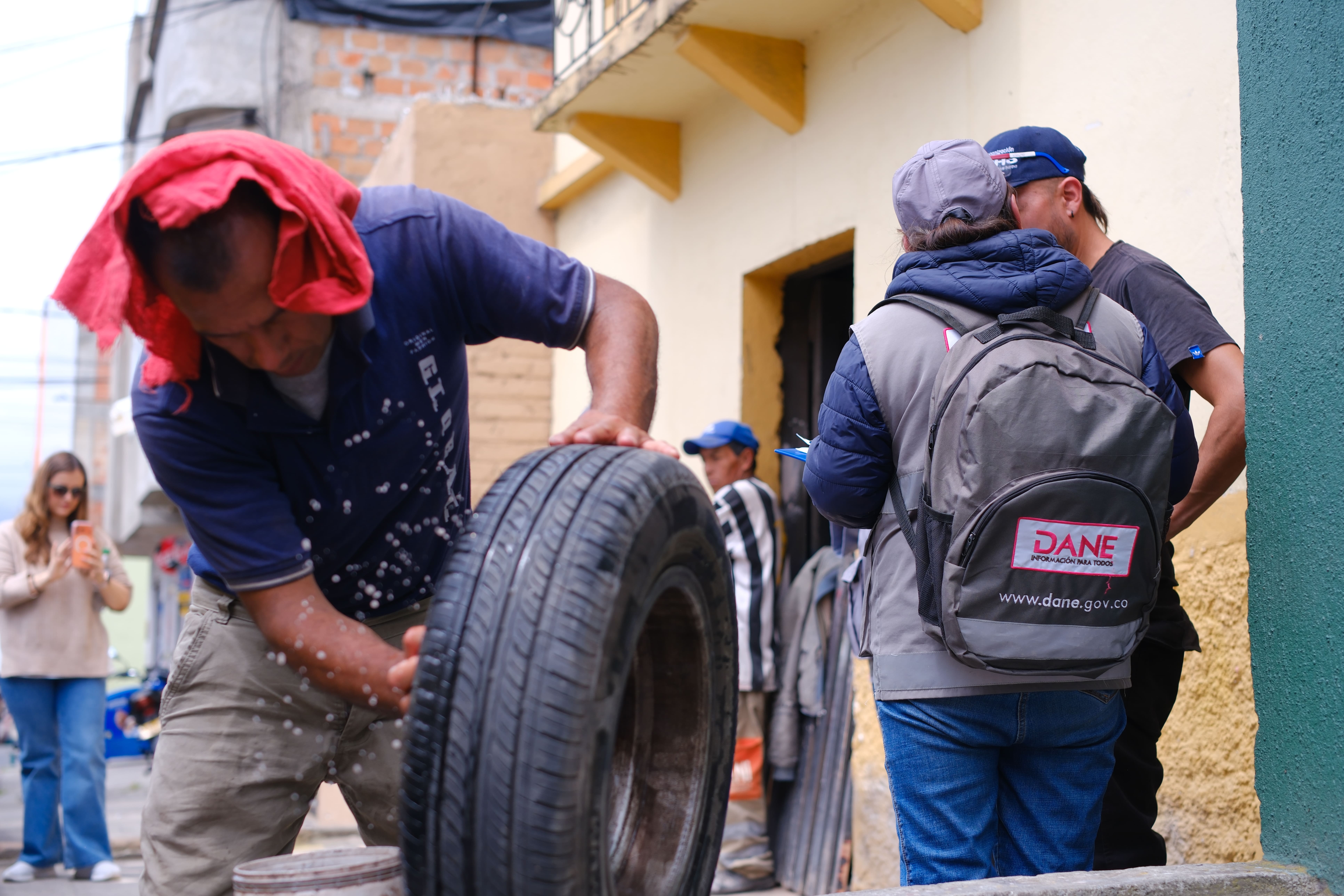 En primer plano se ve un hombre que arregla una llanta en la calle, mientras al lado derecho atrás, un censista del DANE con maleta institucional, entrevista a otra persona