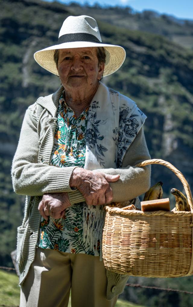Mujer mayor con sombrero blanco de ala ancha sostiene una canasta de mimbre con alimentos, con montañas verdes al fondo.