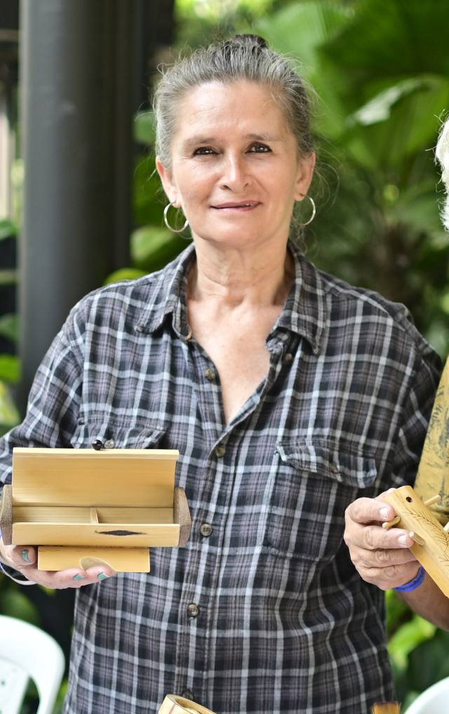 Mujer mayor sostiene cajas y objetos tallados en madera en un evento artesanal, con vegetación verde al fondo.