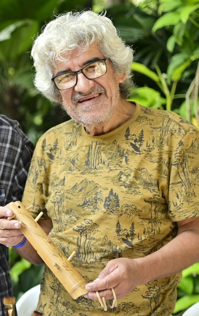 Hombre mayor con gafas y cabello blanco rizado muestra con orgullo una flauta tallada en madera en un evento artesanal.