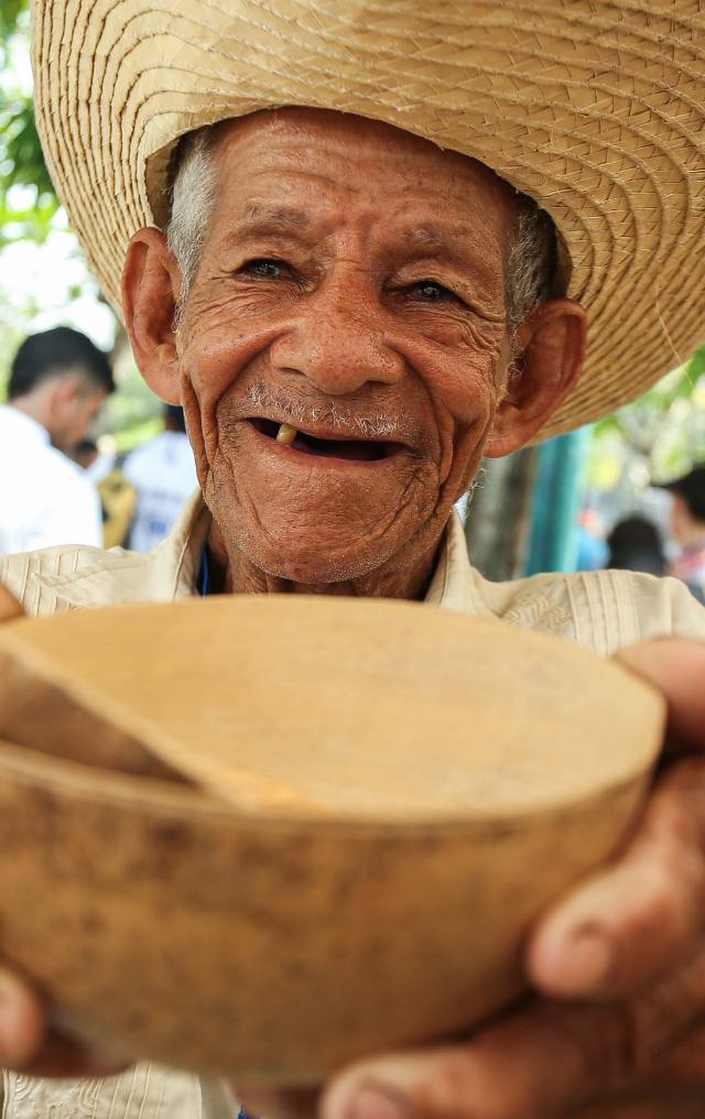 Hombre mayor con sombrero de paja sonríe mientras muestra una totuma tallada en madera hacia la cámara.
