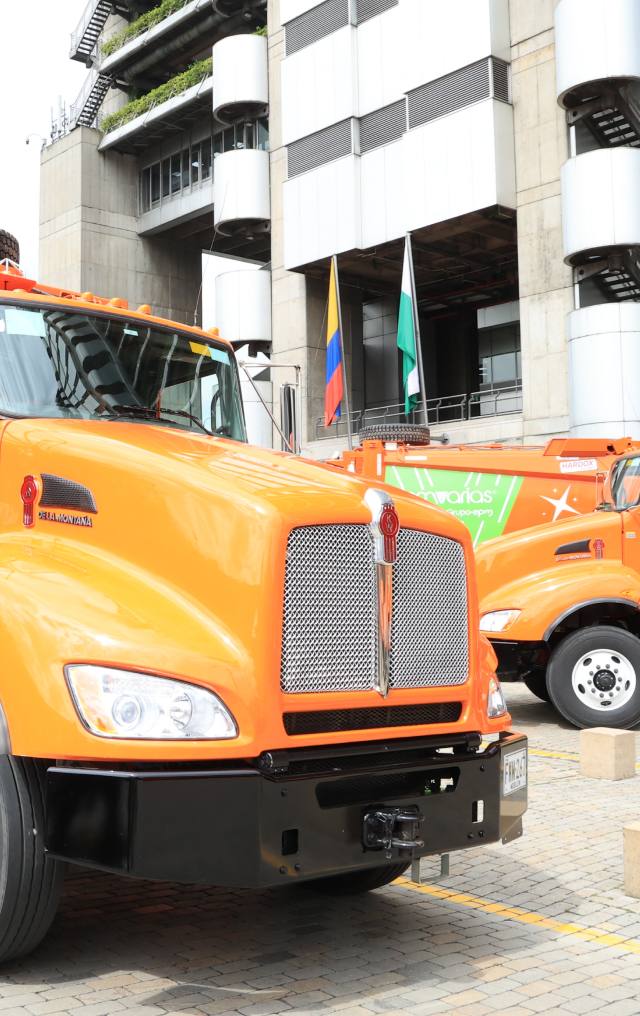 Dos camiones naranjas institucionales estacionados frente a un edificio moderno con banderas de Colombia al fondo.