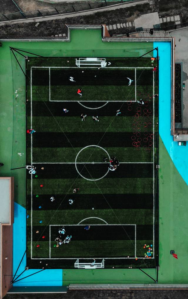 Vista aérea de una cancha de fútbol sintética con franjas verdes y negras, con jugadores en entrenamiento.
