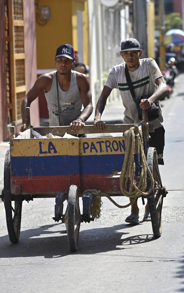 Dos jóvenes empujan una carretilla de madera pintada con los colores de la bandera colombiana por una calle colonial.