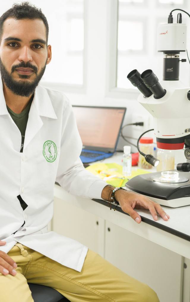 Hombre joven con bata blanca de laboratorio sentado junto a un microscopio binocular en un laboratorio de investigación.