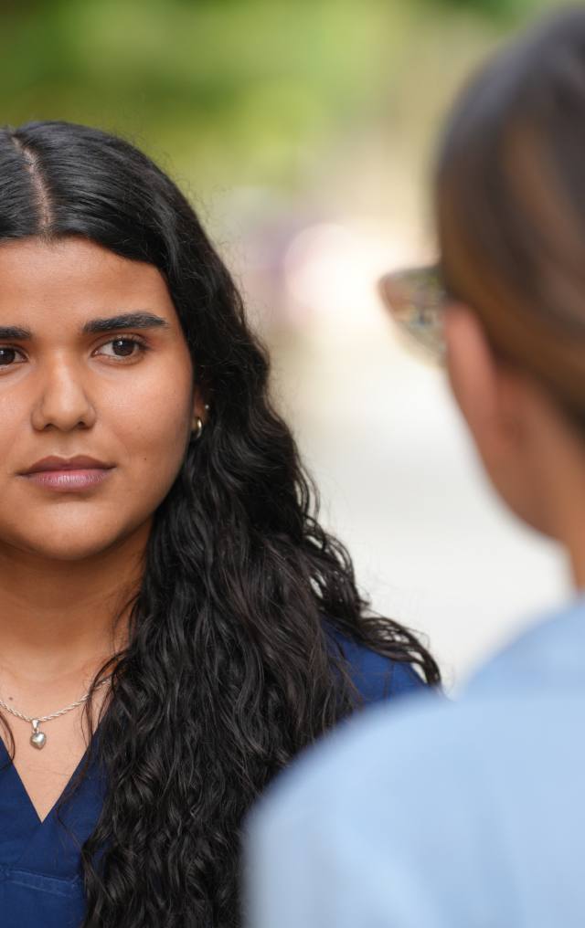 Mujer joven con uniforme azul de salud escucha atentamente a otra persona en un espacio exterior con vegetación.