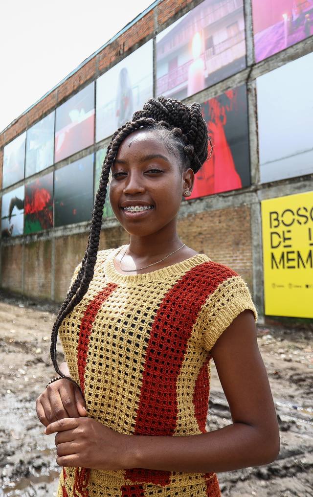 Joven afrodescendiente con trenzas box braids sonríe frente a una pared con fotografías del proyecto Bosque de la Memoria.