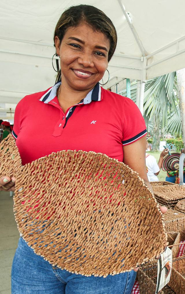 Mujer sonriente con camiseta roja sostiene un canasto tejido en fibra natural en un puesto de mercado artesanal.