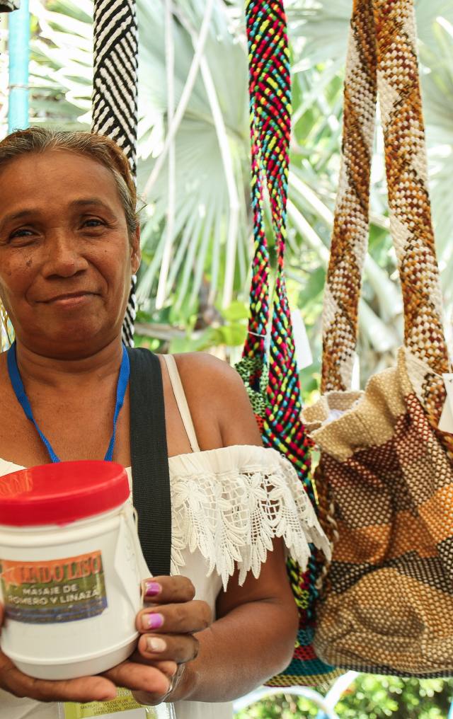 Mujer sonriente sostiene un frasco de crema artesanal rodeada de mochilas y cordones trenzados de colores.