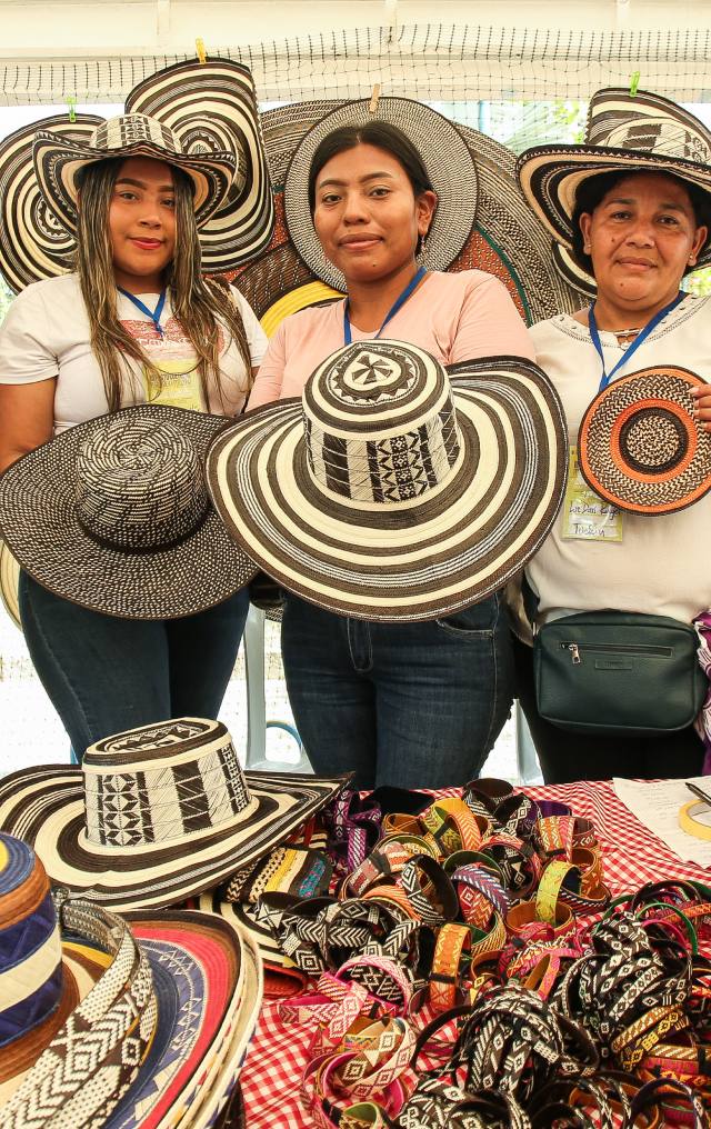 Tres mujeres exhiben sombreros vueltiao tejidos en blanco y negro, junto con otros tejidos, en un puesto de artesanías.