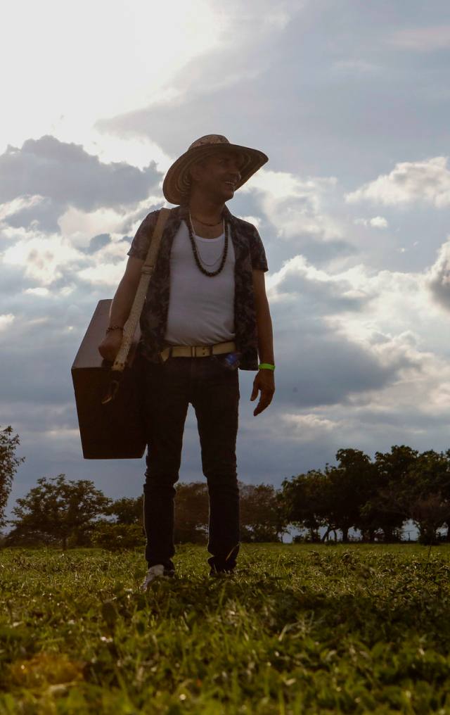 Hombre con sombrero y guitarra cargada al hombro camina sonriente en un campo abierto al atardecer con cielo nublado.