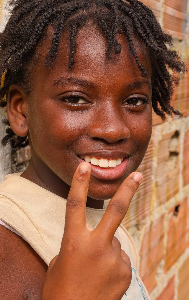 Niña afrodescendiente sonríe con la mano cerca del rostro, apoyada en una pared de ladrillo.