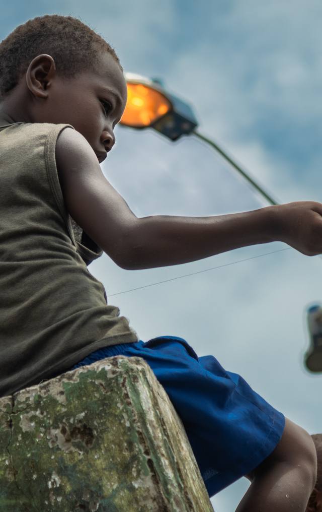 Niño afrodescendiente sentado en un bloque de concreto, mirando hacia abajo con poste de luz encendido al fondo.