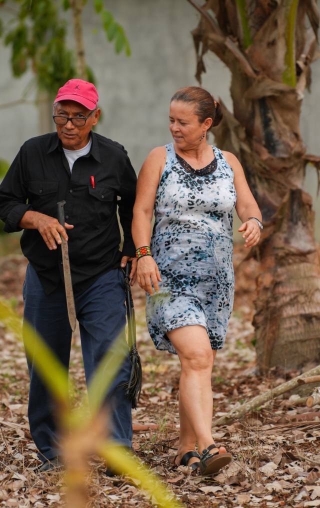 Pareja de adultos camina entre árboles en una finca; el hombre lleva machete y gorra rosada, la mujer viste vestido estampado.