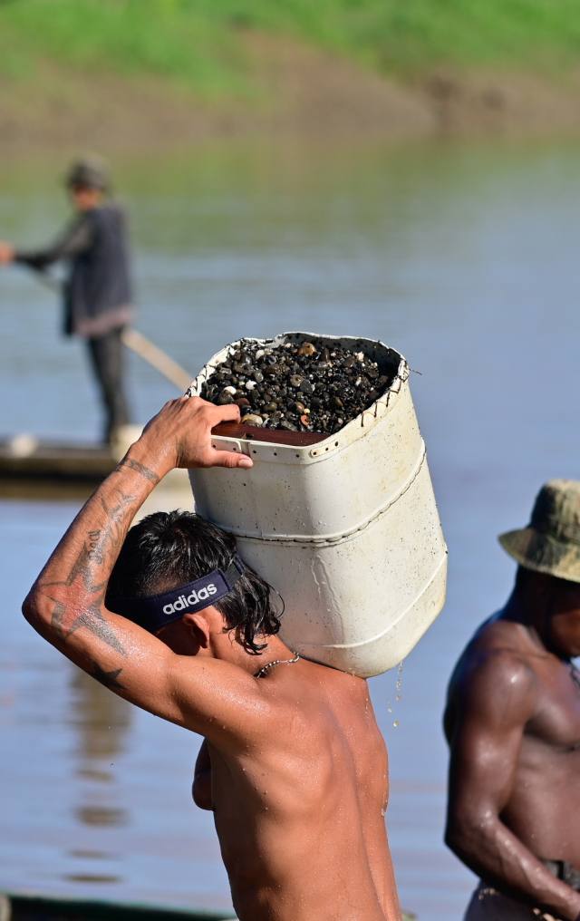 Trabajador carga un balde con material de extracción sobre la cabeza en la orilla de un río, con otra persona en canoa al fondo.