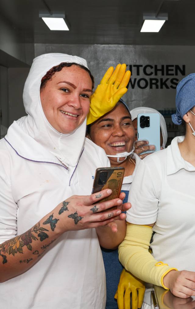 Tres mujeres trabajadoras con uniformes blancos y guantes amarillos se toman una selfie riendo en una cocina industrial.
