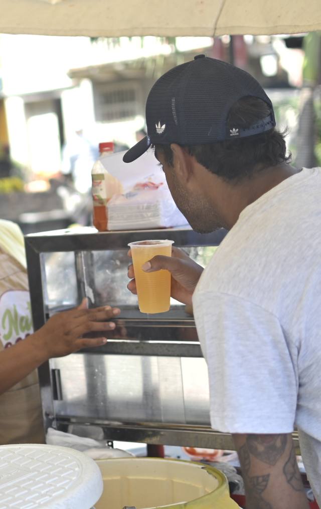 Hombre de espaldas sostiene un vaso de jugo de naranja en un puesto de comida callejera bajo una sombrilla.