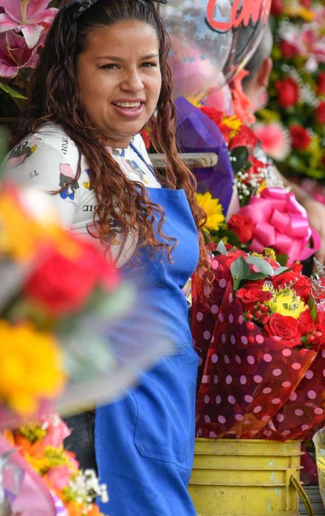 Mujer joven sonriente con delantal azul rodeada de arreglos florales coloridos en un puesto de venta de flores.