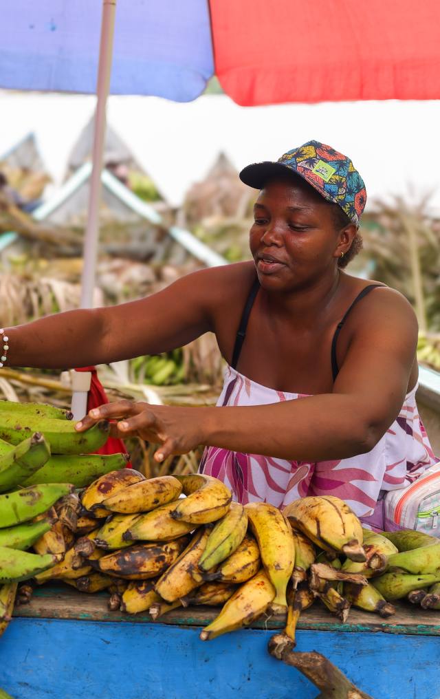 Mujer afrodescendiente organiza plátanos maduros y verdes en un puesto de mercado bajo sombrillas de colores.