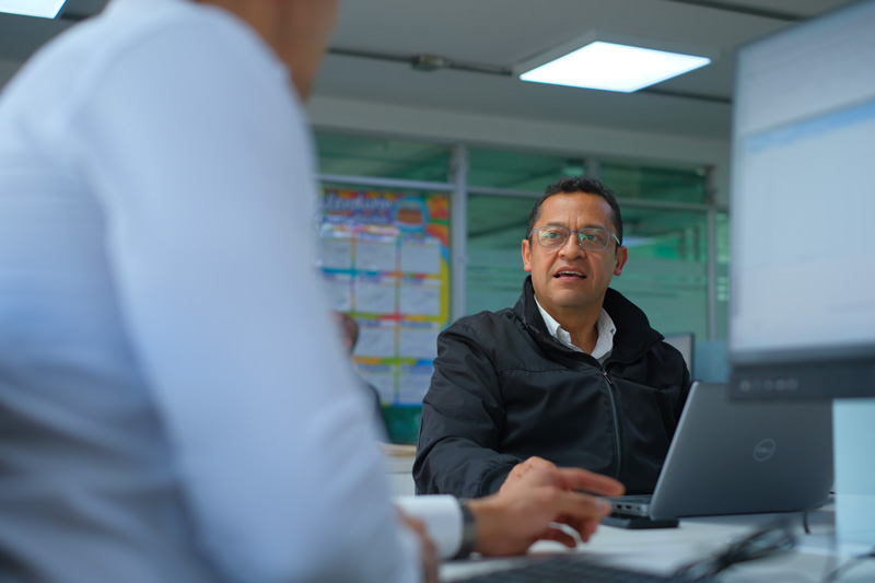 Hombre con gafas y chaqueta negra conversando atentamente con otra persona desde su lugar de trabajo frente a un computador