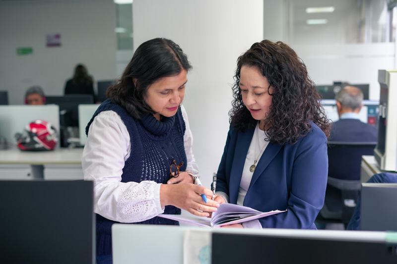Dos mujeres de pie en una oficina, revisando juntas la información de una carpeta de documentos