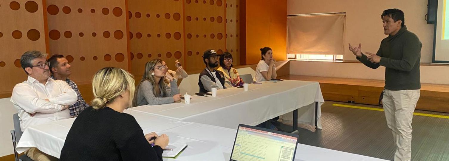 Hombre dando una charla a un grupo de personas sentadas alrededor de mesas blancas en forma de U